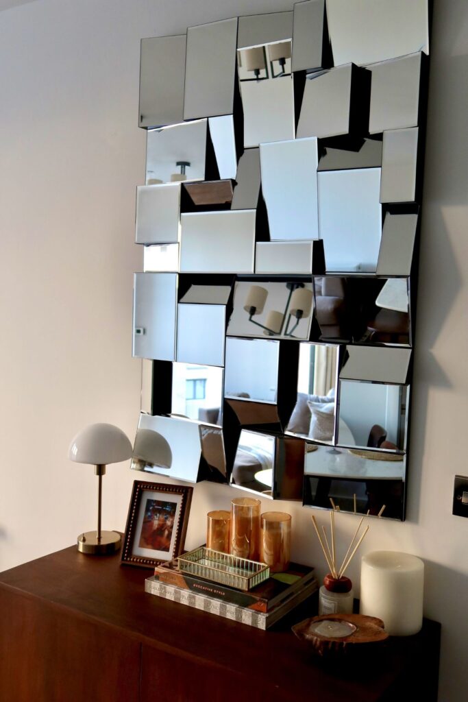 Neutral-toned acacia wood and marble sideboard with square mid century mirror and diffused morning light, with candles and coffee table books, and antique brass lamp.